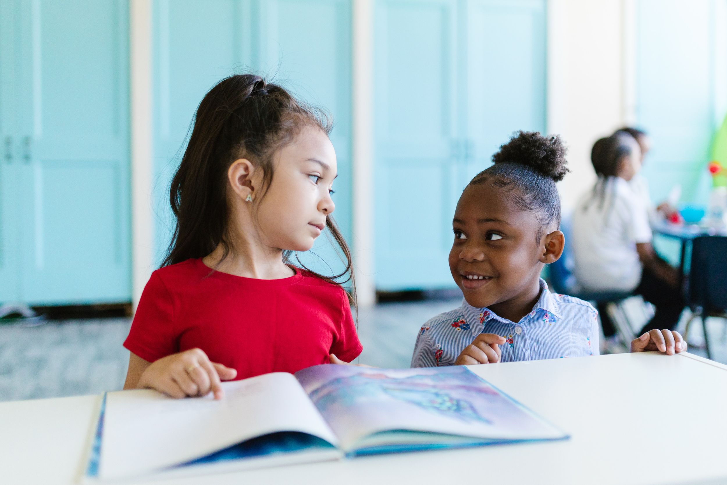 Two little girls stand together and read a book in class.