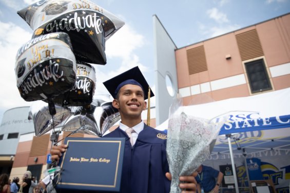 Young graduate in a cap and gown, smilling and holding balloons after IRSC's spring ceremony.