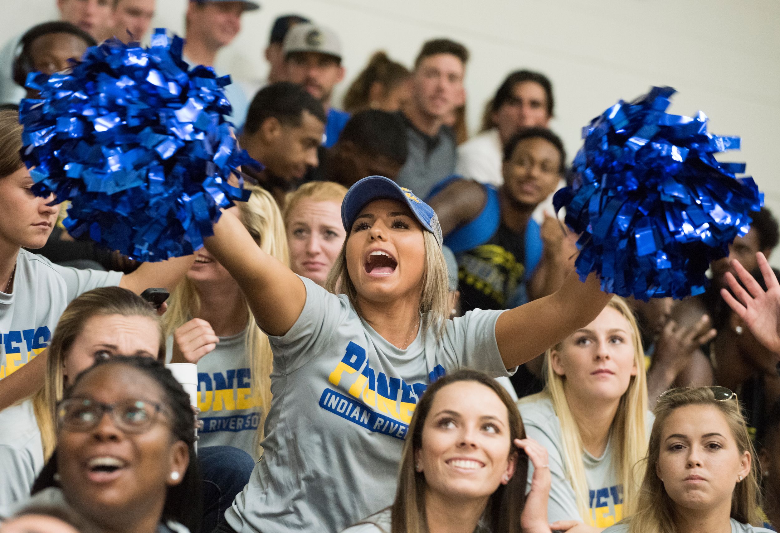 Student cheering in crowded stadium stands with blue pom poms.